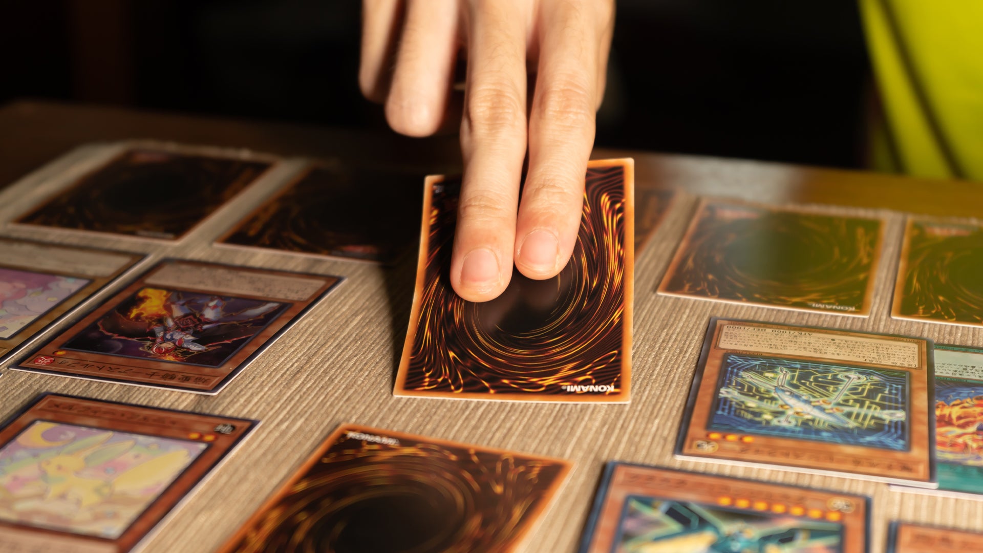 A hand holds a Yu-Gi-Oh! card facedown with two fingers stretched across its back. There are additional Yu-Gi-Oh! cards on the table.