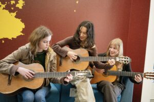 Hanna Gohlke, Shai Eastman, and Vivian BEll practice guitar in the A building lobby.