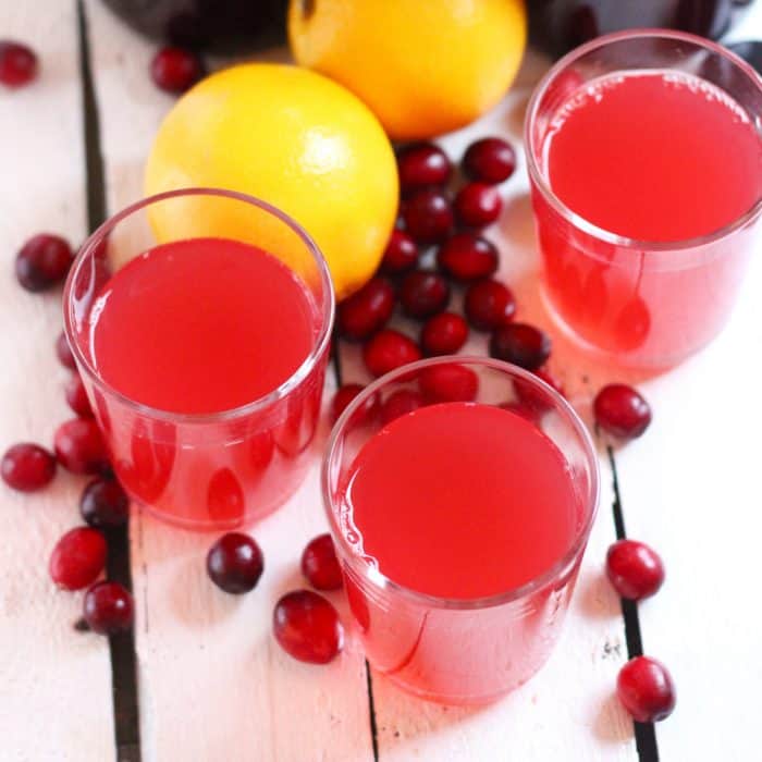 three glasses with cranberry kombucha on white wooden table with fresh cranberries and oranges