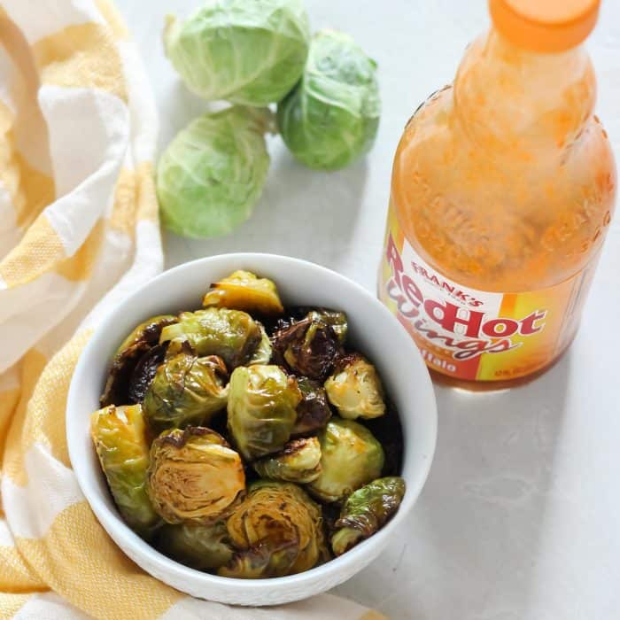 white bowl with roasted Brussels sprouts covered in buffalo sauce, fresh Brussels sprouts, bottle of buffalo sauce, yellow and white striped dish towel