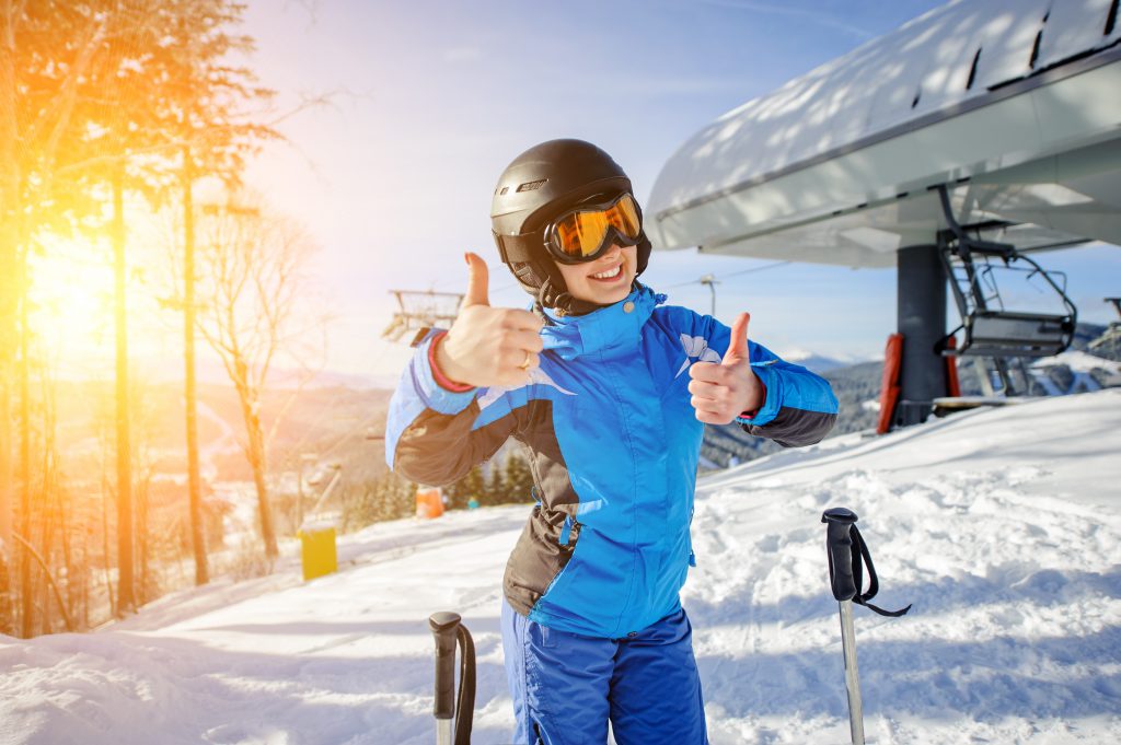 Portrait of young female skier at ski resort smiling and showing thumbs up. Winter sports concept. Woman is wearing blue jacket and blue pants, helmet and orange goggles.