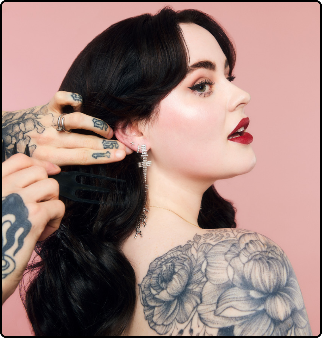 Isabelle Kate wearing red lipstick and black eyeliner, looking over her shoulder. Someone's tattooed hands are holding her hair back, revealing a set of sparkling earrings.