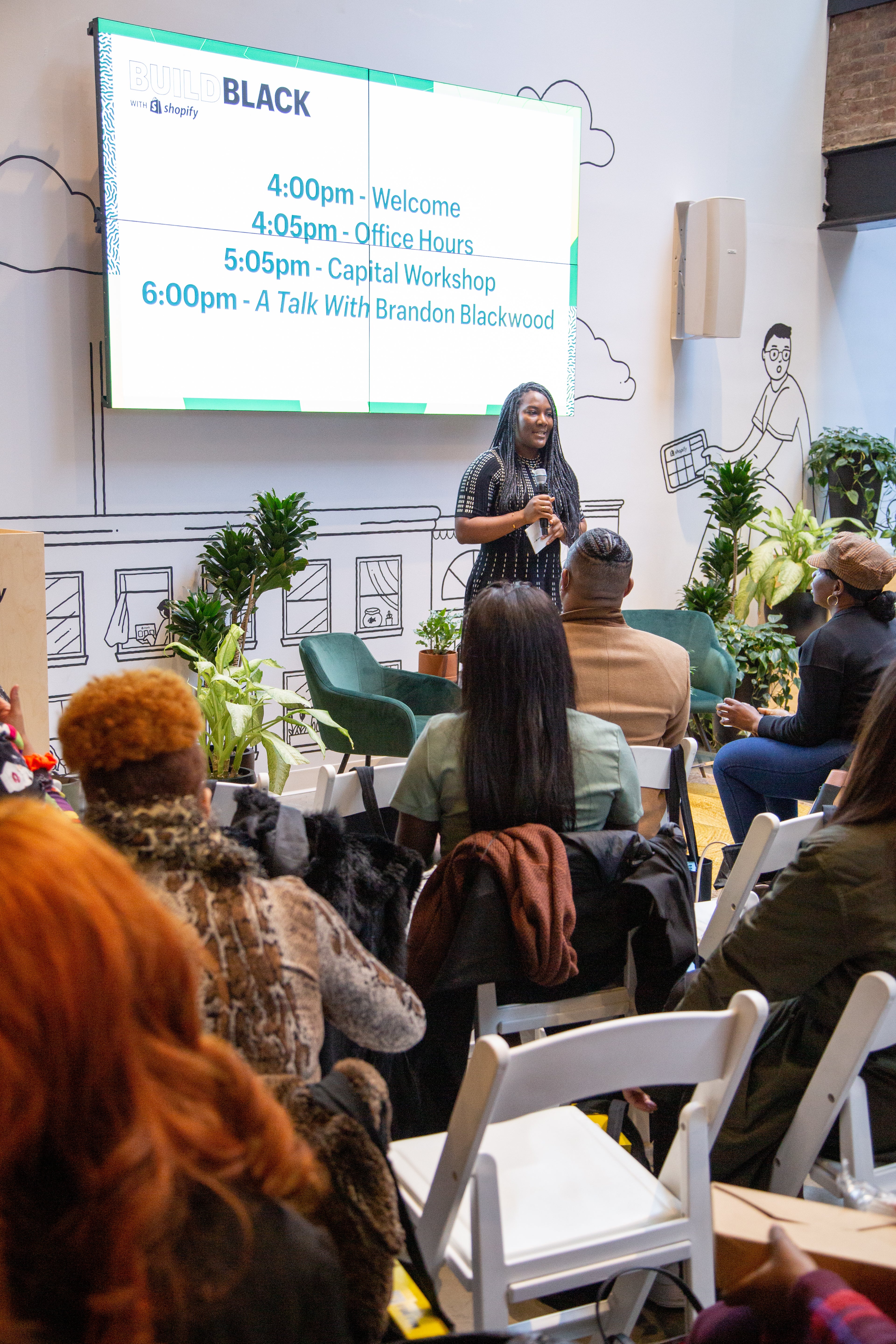 A photo of the Build Black program lead, holding a mic infront of an audience of seated guests at a Build Black event. There's a large monitor behind the program lead indicating the schedule for the event.