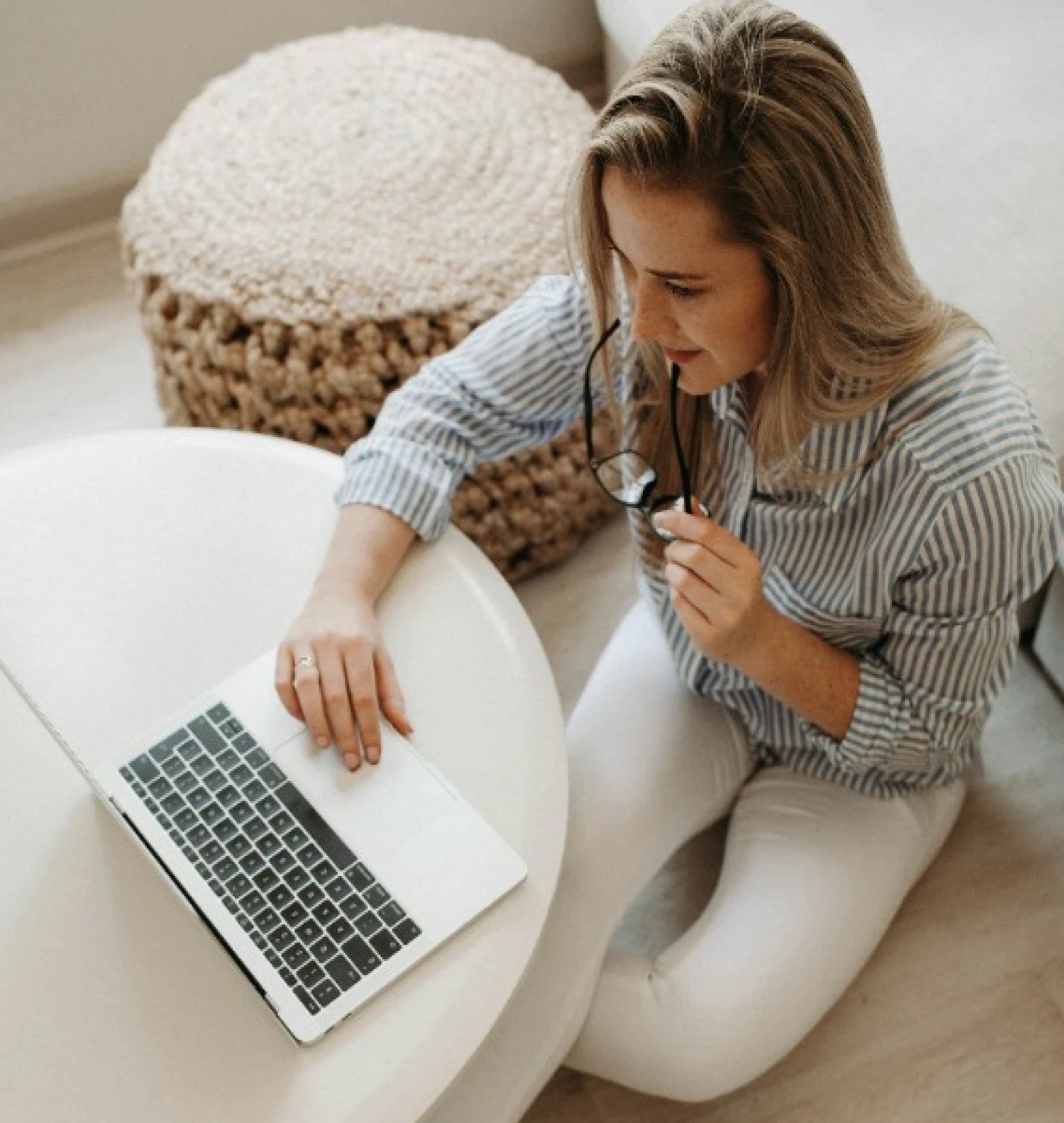 Woman sitting and working on a laptop, holding glasses in hand.