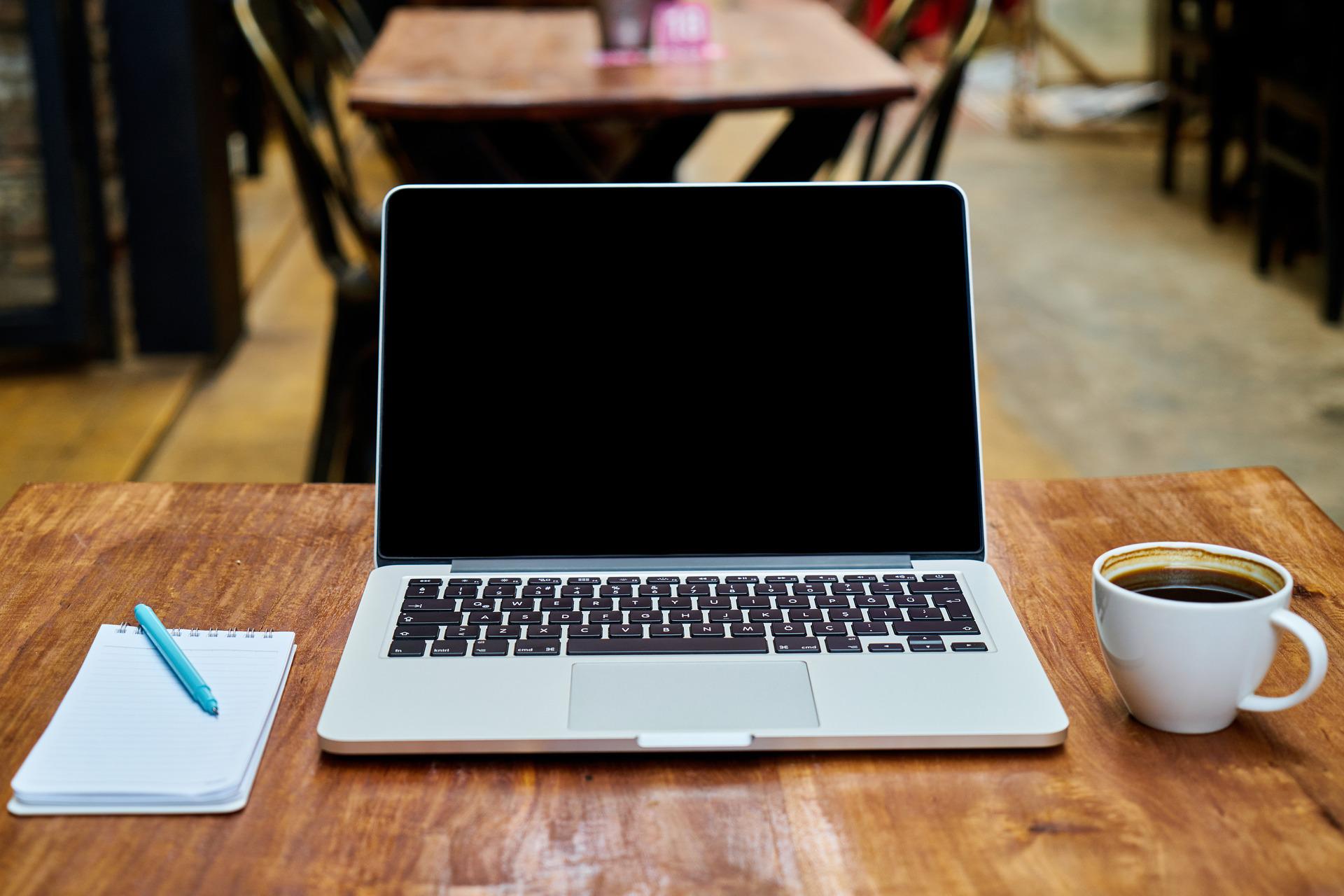 Auf einem Schreibtisch aus Holz steht ein aufgeschlagener, ausgeschalteter Laptop neben einer gefüllten Kaffeetasse rechts und einem Notizbuch und Stift links.