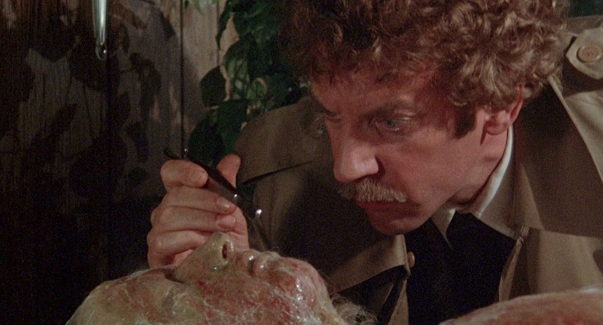 A man (Donald Sutherland) examines the face of a body enmeshed in a strange web-like skin of sinuous fibers.