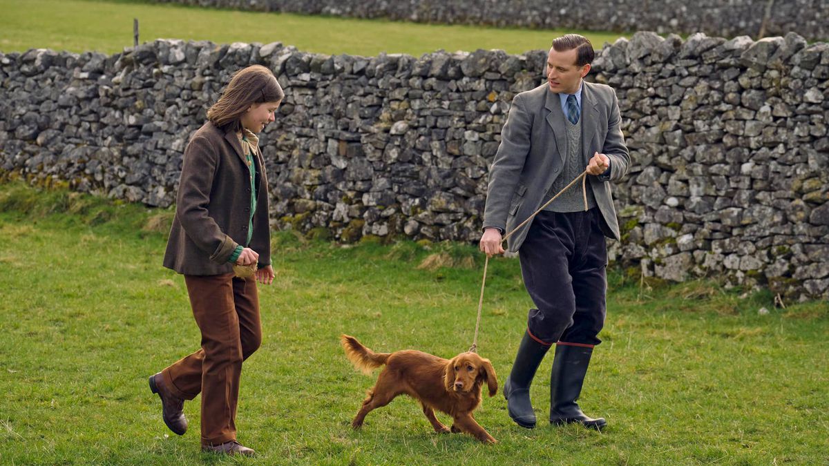 James Herriot, wearing a grey coat and blue trousers, walks a small dog on a rope leash next to a child.