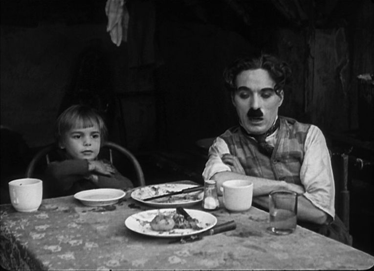 Charlie Chaplin and Jackie Coogan sit with their arms crossed at a dinner table in The Kid