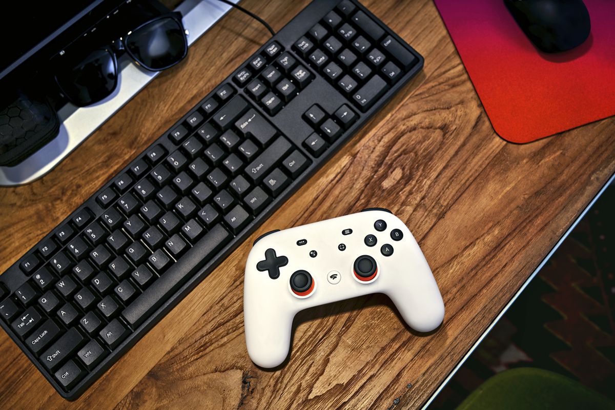 A Google Stadia controller rests on a wood desk next to a black computer keyboard