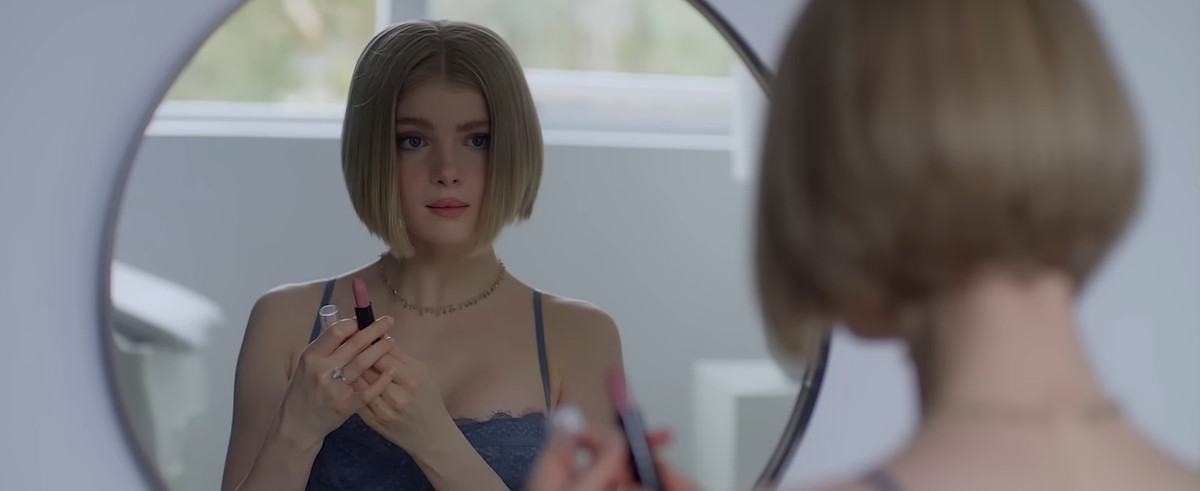 Meredith (Elena Kampouris) examines herself in a mirror while holding up lipstick in Wifelike