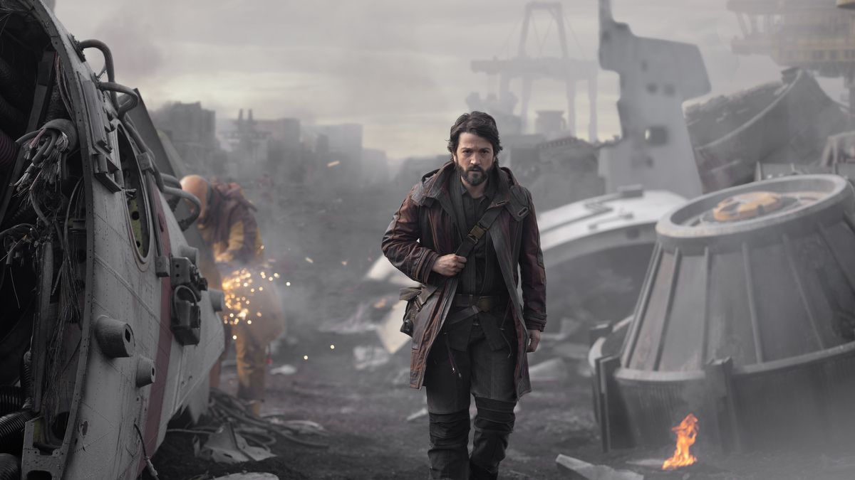 Diego Luna as Cassian Andor walks through a field of scrap, with machines broken and on fire.