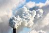 A photograph of the top of a factory chimney shown against the sky, emitting a dense smoke into the air