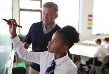 A photograph of a secondary or high school student writing on a whiteboard, while her teacher provides feedback