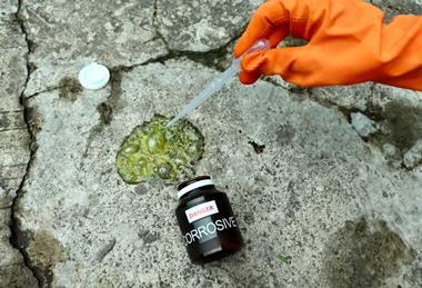 A photograph showing a bottle of acid spilled on a rock surface; a gloved hand holds a pipette over the spillage