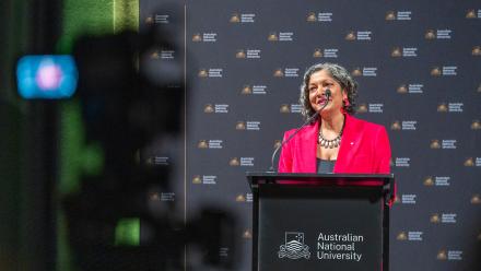 A woman with short black hair wearing a pink blazer speaks at a lectern. 