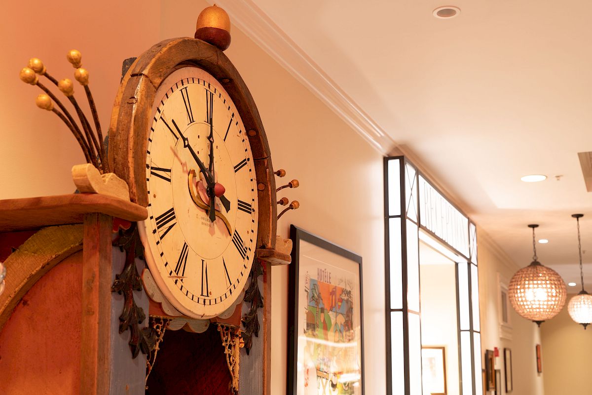 The image shows a decorative clock with Roman numerals, positioned in a well-lit hallway with framed pictures and modern ceiling lights.