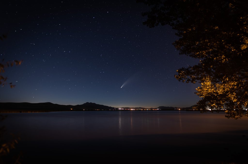 A serene night sky is filled with stars and features a visible comet with a luminous tail. A calm body of water reflects the starlight and city lights in the distance. Trees frame the scene on the right, with silhouettes of mountains on the horizon.