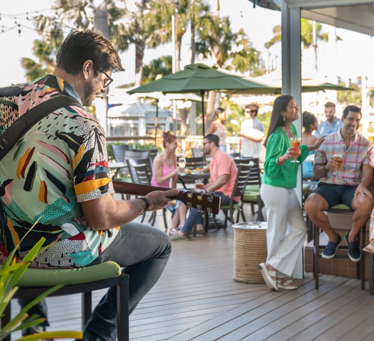 A Person Playing Guitar In Front Of A Group Of People Sitting At Tables