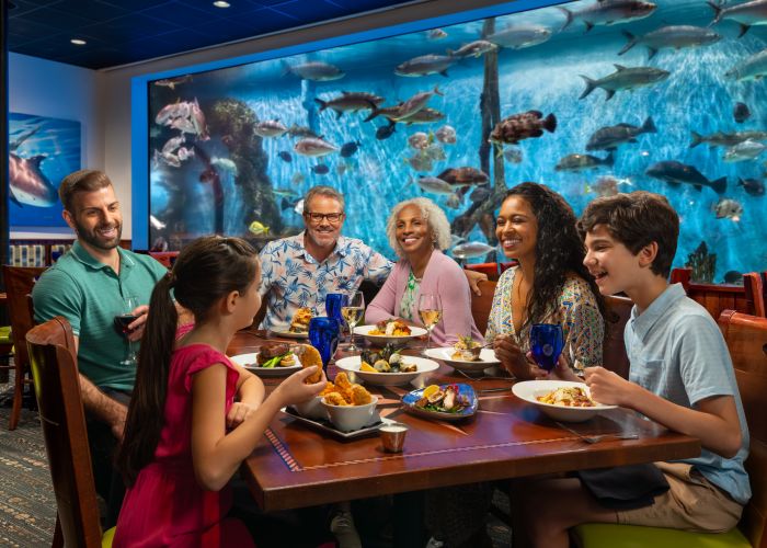 A Group Of People Sitting At A Table With Food And Fish In The Background