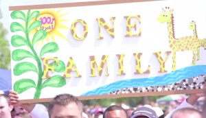 Protesters at a Market Basket rally hold a sign saying "One Family," featuring giraffes, the mascot they've adopted. NewsHour still image.