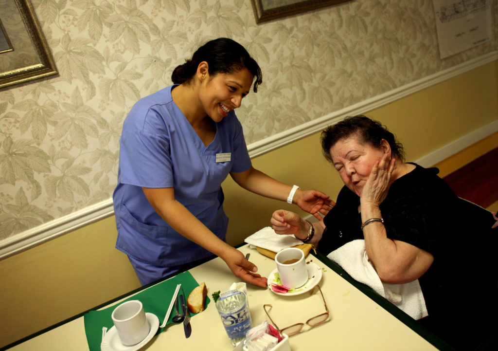 Wendy Estrada, a 30-year-old Honduran immigrant training to become a certified nursing assistant listens to Center at Park West resident Maria Ruvalcaba sing. Immigrants make up a large share of health care aides in the U.S. Photo by Barbara Davidson/Los Angeles Times via Getty Images