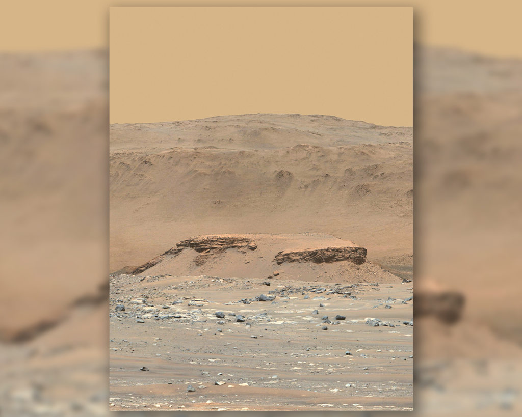 A prominent layered mound named Kodiak stands approximately 2 km from the Perseverance rover and is a remnant of the ancient Jezero delta. In this enhanced color image, the mesa-like rock appears a light rust color, while the rocks below it have a bluish tint, and the edges of Jezero Crater loom tall in the backdrop before a beige Martian sky