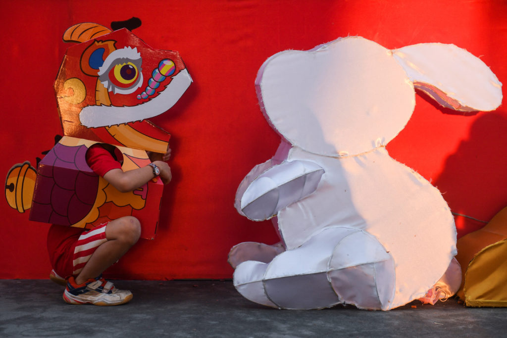 A boy, wearing a paper lion dance head, sits next to a white rabbit lantern on the first day of the Lunar New Year of the ...