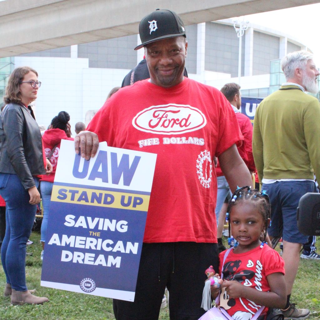 Edward Fife, a member of UAW Local 600 who works at Ford Motor Company’s Dearborn Truck Plant, attended the UAW’s “Stand up for our Communities against Corporate Greed” rally in Detroit with his family. He is holding a sign that reads "UAW Stand Up Saving the American Dream. September 15, 2023. Photo by Frances Kai-Hwa Wang for the PBS NewsHour