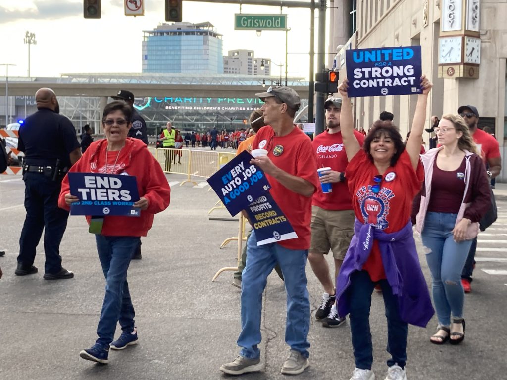 UAW members march past the North American International Detroit Auto Show to Blue Cross Blue Shield to show support for other UAW members who are also striking after the UAW’s “Stand up for our Communities against Corporate Greed” rally in Detroit while holding signs that say “End tiers no second class workers,” “Every auto job a good job,” “Record profits record contracts,” and “United for a strong contract.” September 15, 2023. Photo by Frances Kai-Hwa Wang for the PBS NewsHour