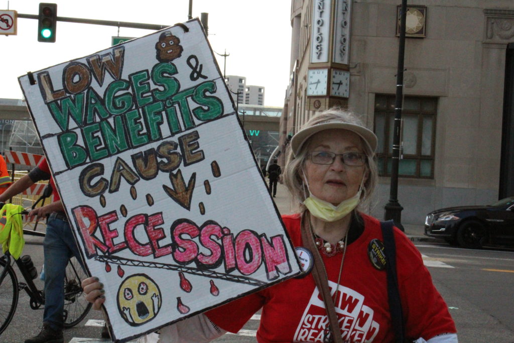 Judy Wraight, a retired autoworker with 34 years seniority at Ford’s River Rouge Complex in Dearborn and a member of UAW Local 600, pickets with her homemade sign, “low wages and benefits cause recession,” after the UAW’s “Stand up for our Communities against Corporate Greed” rally in Detroit. September 15, 2023. Photo by Frances Kai-Hwa Wang for the PBS NewsHour