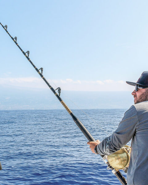 a man faces away from the camera so only his profile shows. He is wearing a gray long sleeved shirt, baseball cap and sunglasses. He is holding a large fishing rod pointed towards the ocean