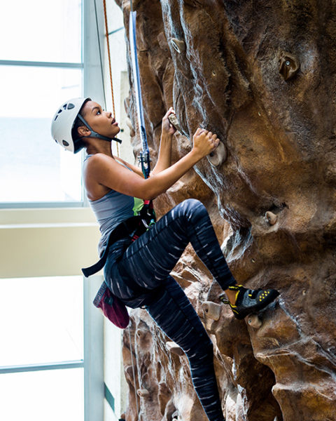 a woman wearing a white helmet, blue tank top and striped leggings climbs up the rock wall