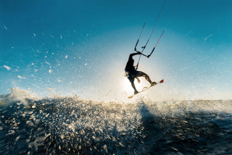 a man on a kite board flies through the air. He grasps on to the bar attached to the kite with one hand. The camera is sprayed with water from the waves he is surfing.