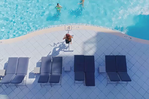 aerial shot of the pool. there is a lifeguard watching two people swim in the pool. There are gray lounge chairs lining the pool