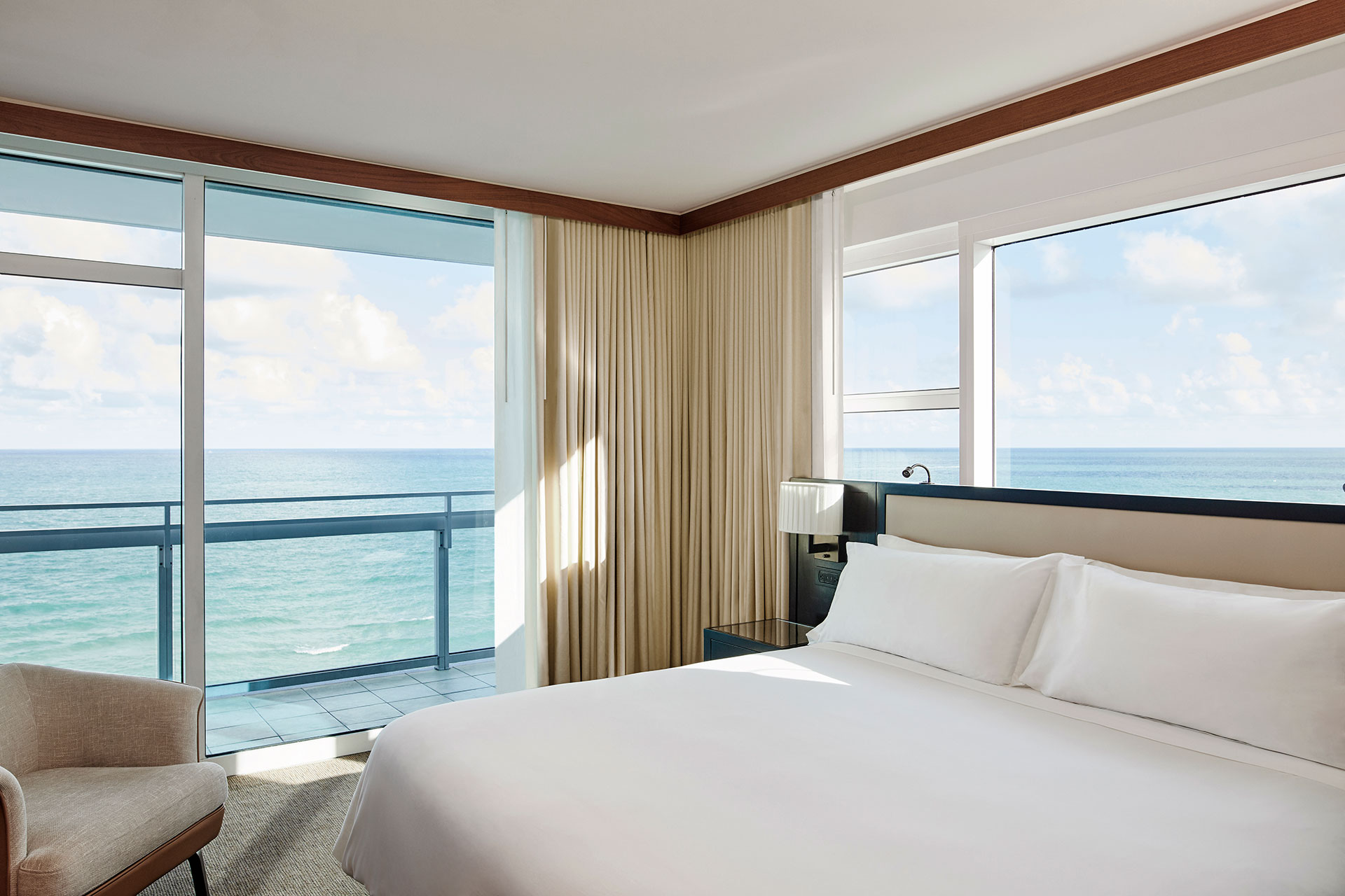 an ocean view suite with a balcony. The bed is on the right side of the room with crisp white linens. There is a black bedside table and a light colored chair in the left corner.