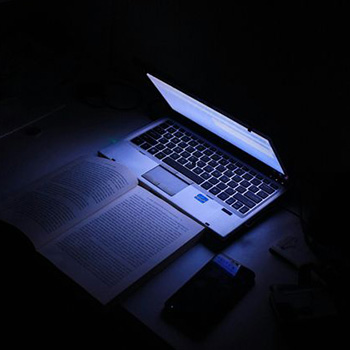 An open laptop casts a purple glow on the textbook in front of it.