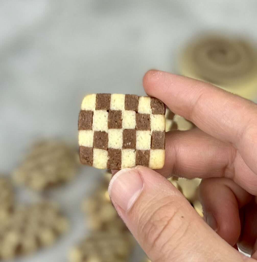 holding a checkerboard cookie in hand above tray of cookies on table 
