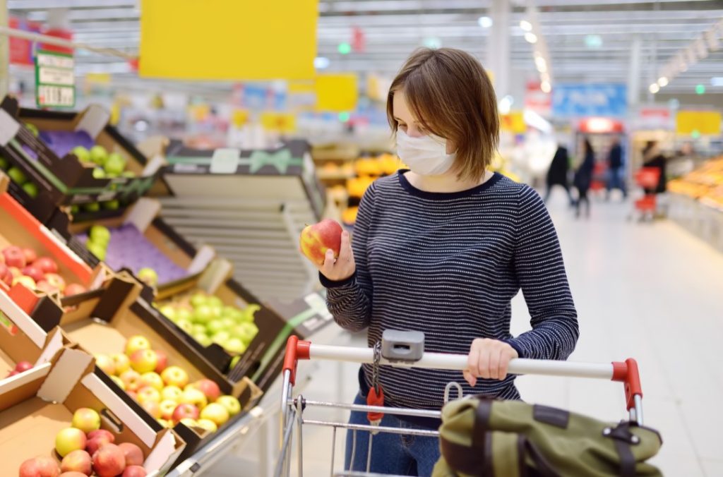 mulher em supermercado escolhendo uma fruta. Ela está usando máscara de proteção