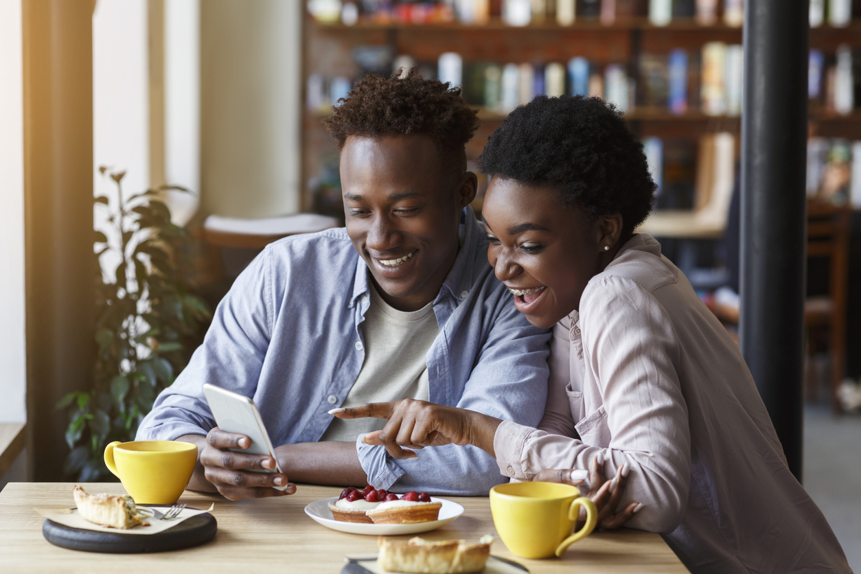 Lindo casal afro-americano com smartphone num encontro em cafeteria, avaliando sobre estratégias de On Trade e Off Trade.
