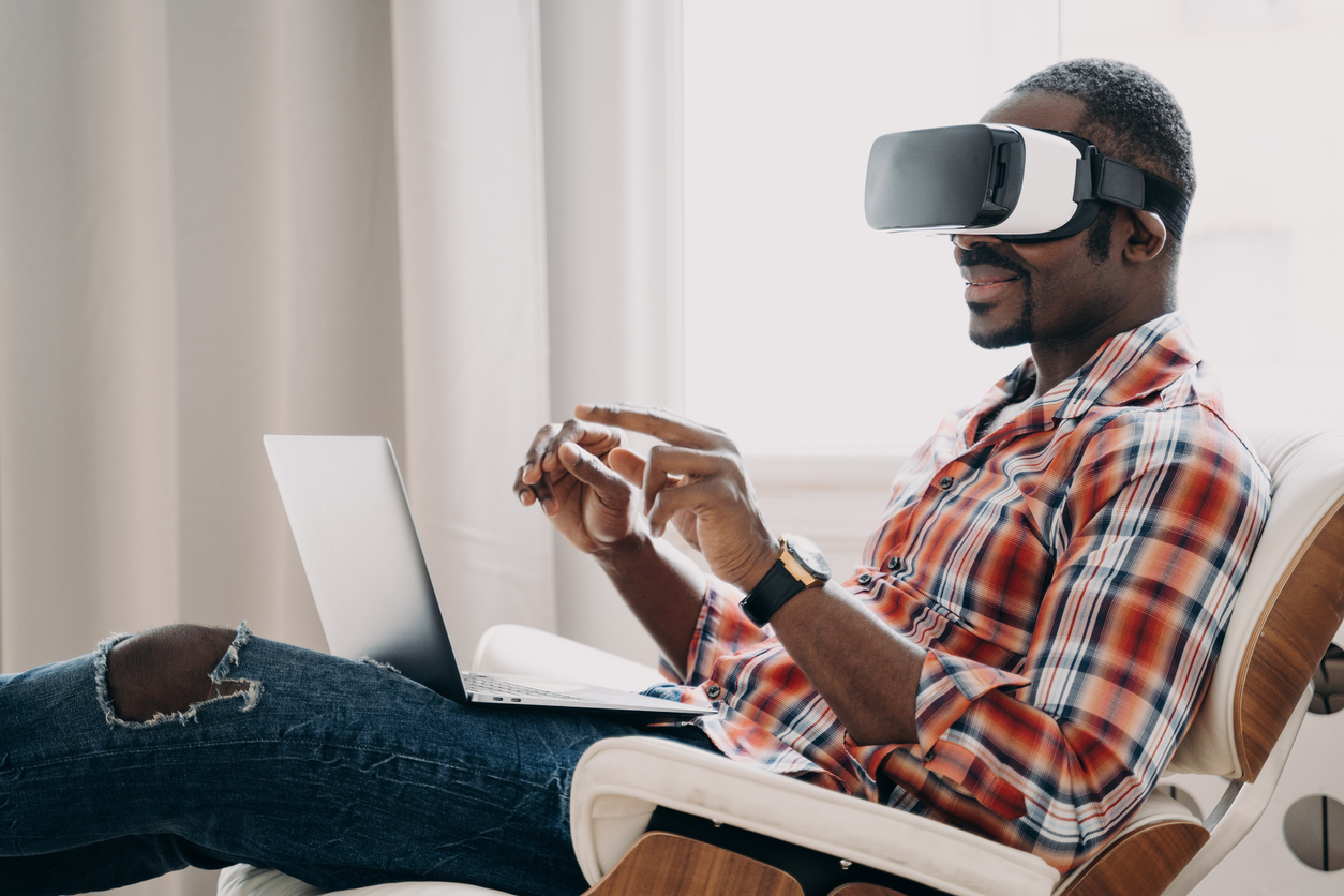 Um homem negro de camisa xadrez vermelha e calça jeans está deitado em uma poltrona carregando um notebook no seu colo. Ele está usando um óculos de inteligência artificial para realizar uma compra em um negócio phygital
