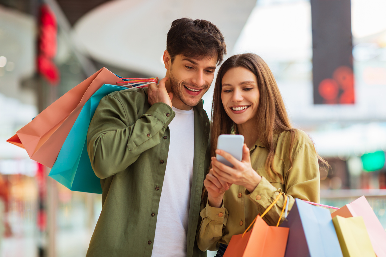 Um casal branco, de cabelo castanho, blusa verde com botões está em um shopping segurando varias sacolas de compra. A mulher está segurando o celular e mostrando para o namorado as várias promoções da Black Friday para eles aproveitarem