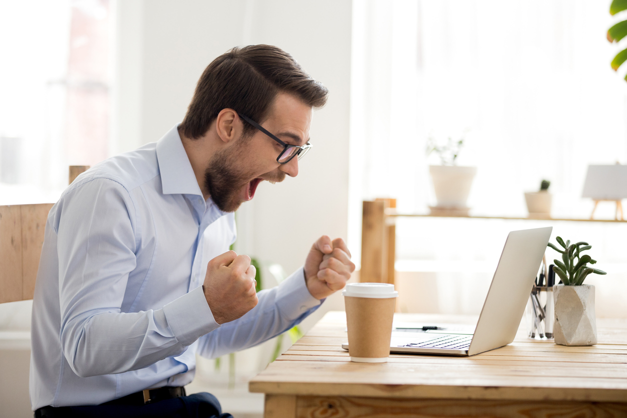 Um homem brano, de cabelo castanho curto e barba curta, está usando uma camisa social azul clara e um óculos de grau com armação preta. Ele está sentado em uma mesa de frente para um notebook e um copo de café. Ele está feliz, comemorando porque obteve sucesso ao aplicar tecnicas para analise de concorrencia
