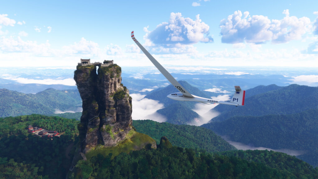 A glider soars over mountainous terrain. There is a large stone pillar visible in the background