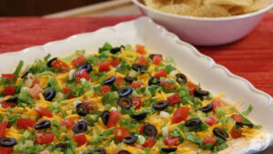 Classic Easy Taco Dip on a serving plate topped with shredded cheddar cheese, lettuce, roma tomatoes, sliced black olives, and green onions.