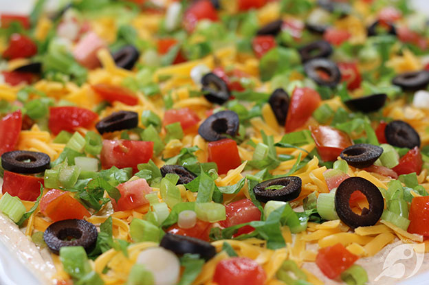 Closeup of Classic Easy Taco Dip on a serving plate topped with shredded cheddar cheese, lettuce, roma tomatoes, sliced black olives, and green onions.