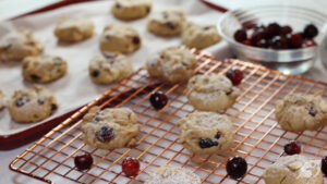 Decadent White Chocolate and Cranberry Oatmeal Cookies with a powdered sugar sprinkle on top. Cooling on a copper wire rack. Background shows a bowl of fresh cranberries and a cookie sheet with baked cookies.