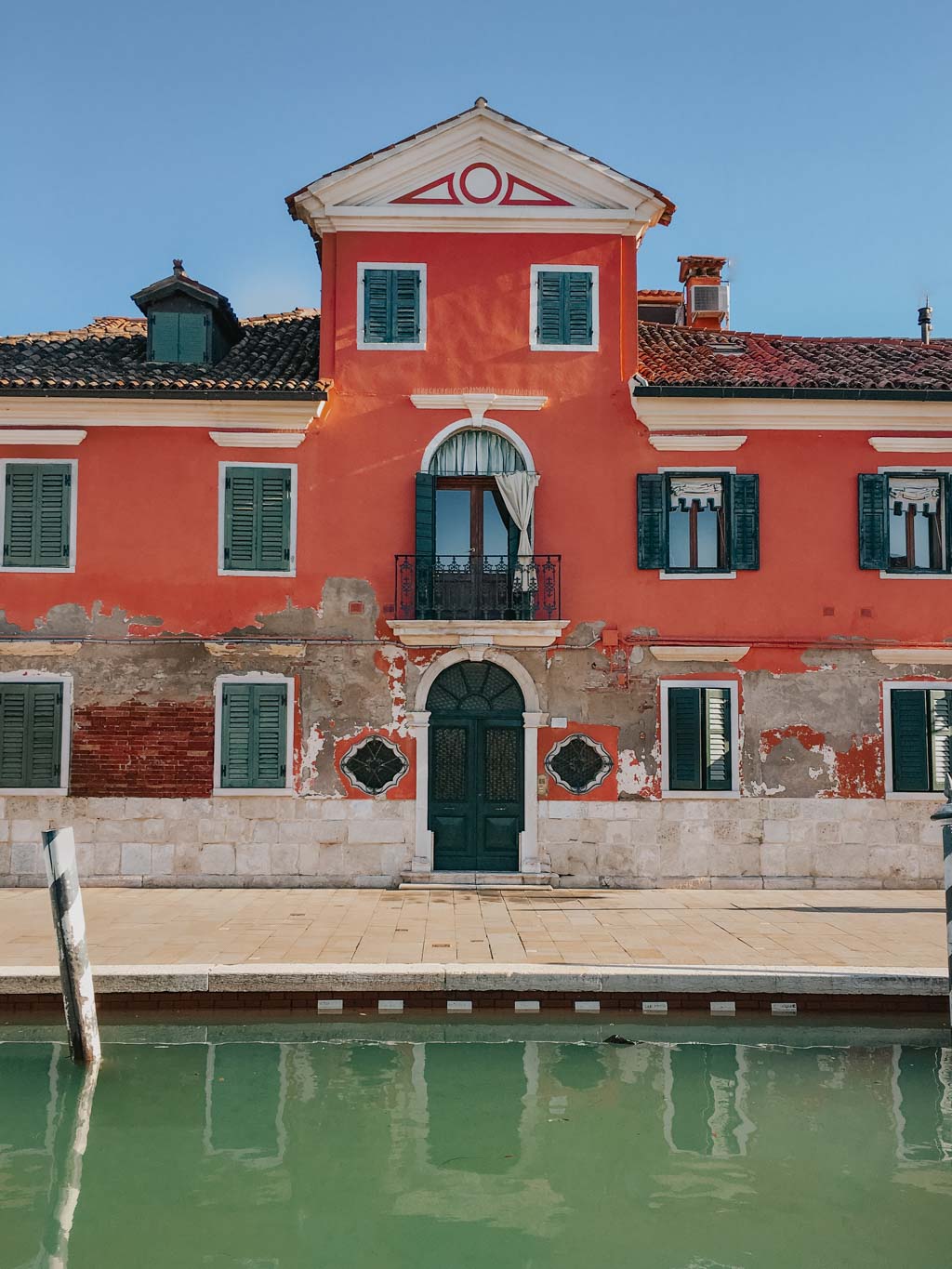 Venice 2 day itinerary will take you to Burano Island with bright red buildings with green window shutters along a water canal on a bright blue sunny day