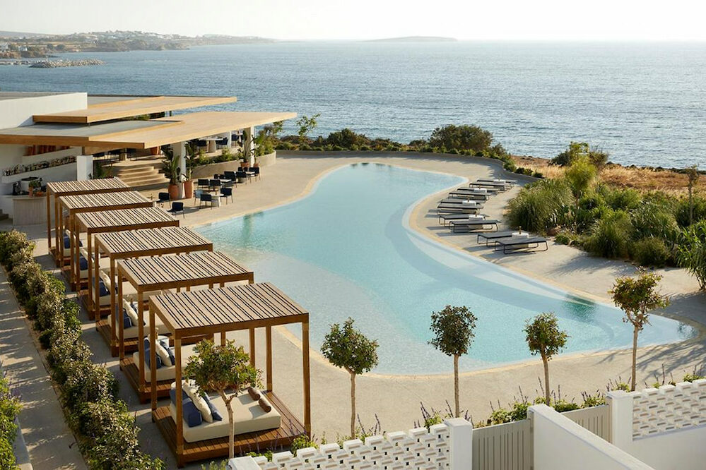 large organic pool surrounded by sun loungers and cabanas and low level boutique hotel in Paros Island Greece