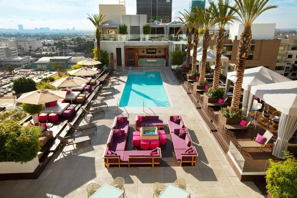 birds eye perspective of a Hollywood hotel rooftop pool and lounge area with pink sun furniture, white umbrellas and potted palm trees