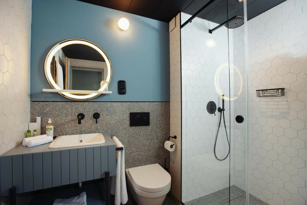 A round mirror with lights hanging in the wall of the bathroom near the shower area